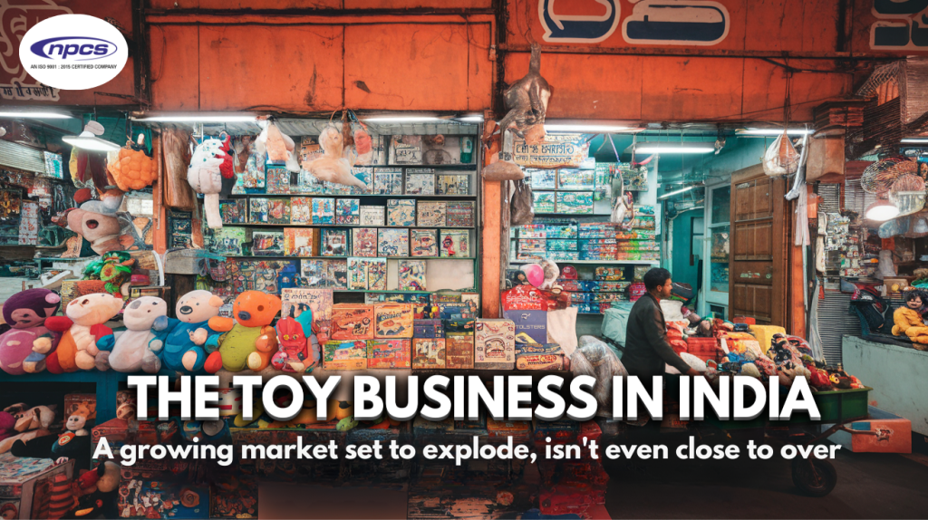 A toy shop in India with colorful stuffed animals, board games, and toy boxes displayed on shelves and hanging from the ceiling. A vendor sits behind a cart filled with toys. The shop has an orange exterior, bright lights, and a bustling market atmosphere. Text overlay reads 'The Toy Business in India: A growing market set to explode, isn't even close to over.' The NPCS logo is visible in the top left corner."